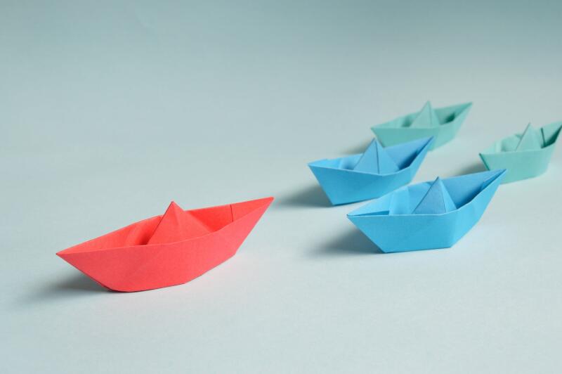 Ein rotes Papierschiff fährt vor mehreren blauen Papierschiffen.